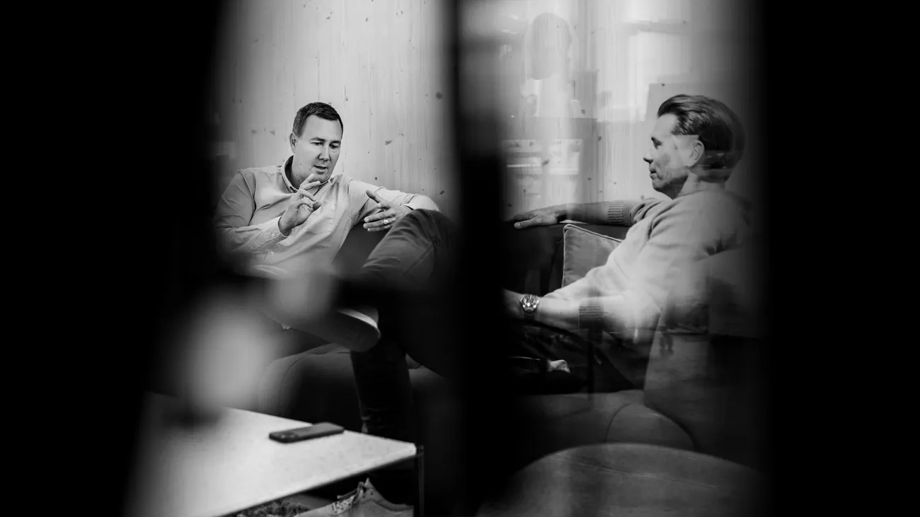 A black-and-white photo of two men talking on a sofa.
